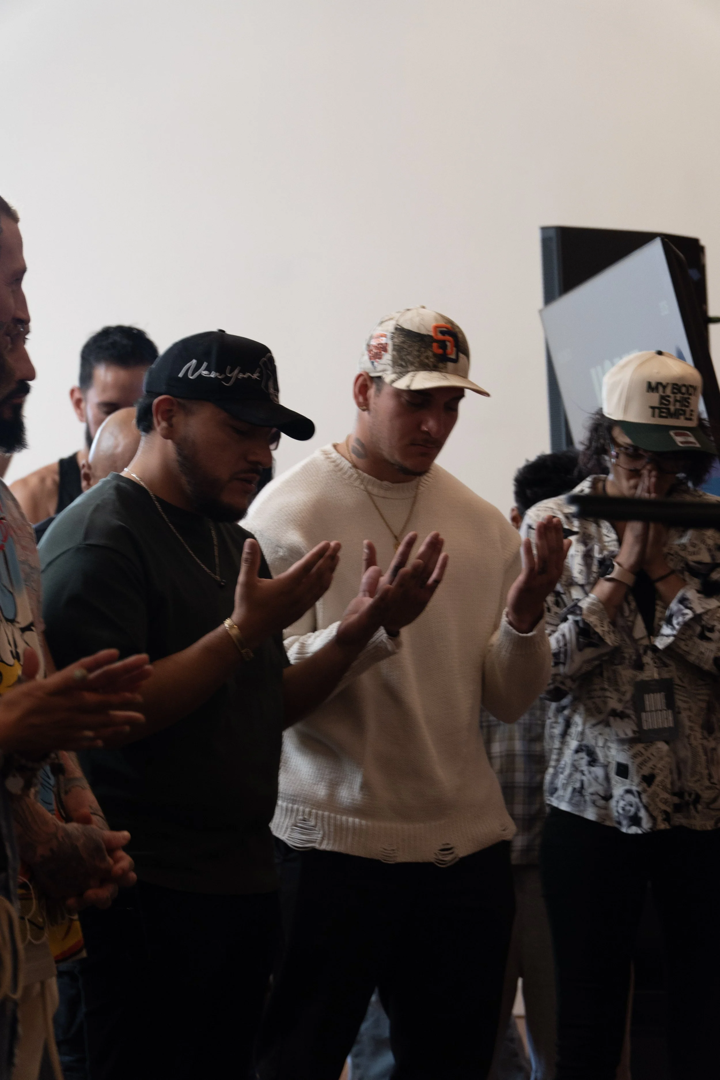 Group of people praying with eyes closed and hands raised in prayer in a room with a plain white wall and a monitor in the background.