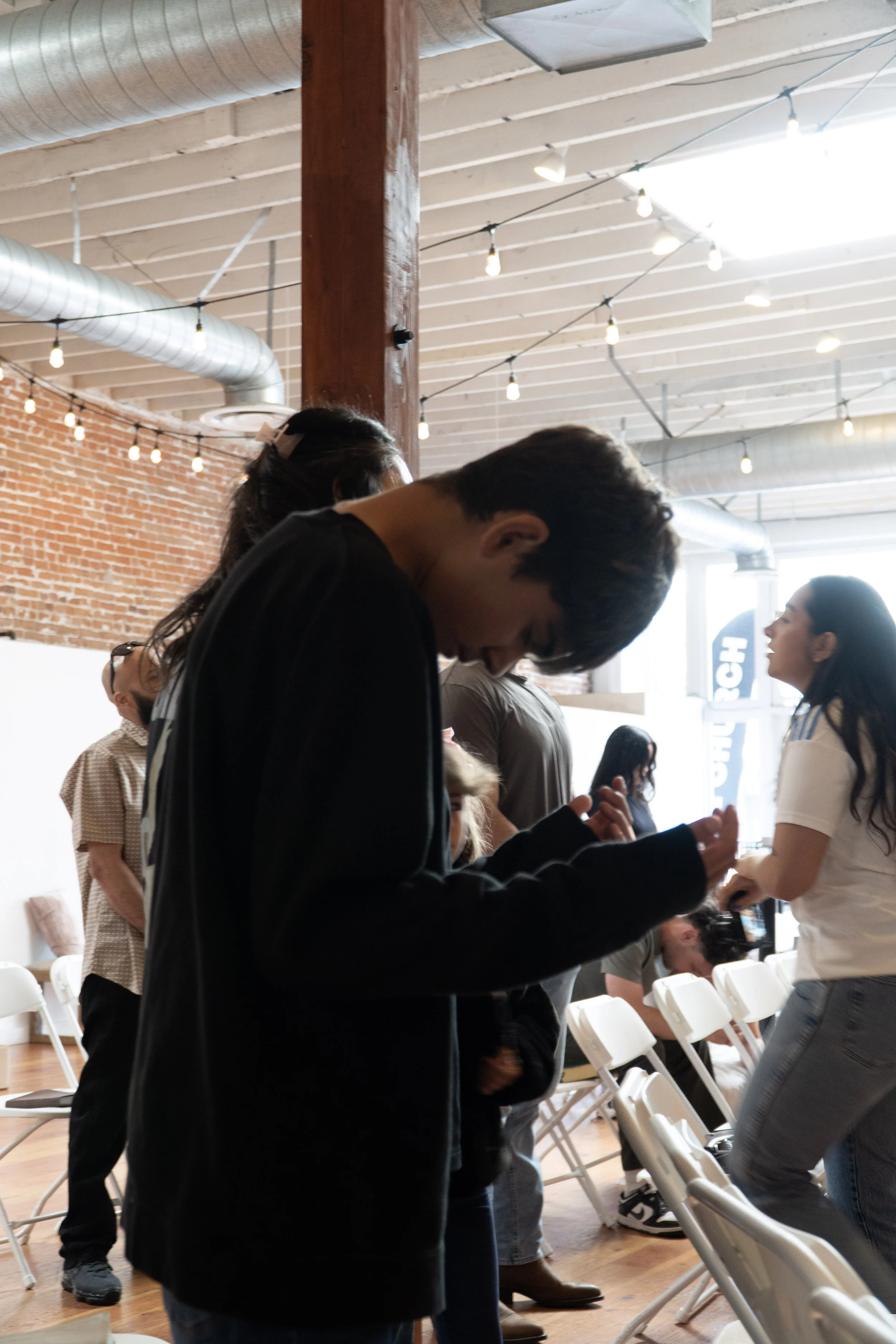 People gathered in a modern indoor space with exposed brick walls, string lights, and large windows, some standing and talking, others sitting on chairs.