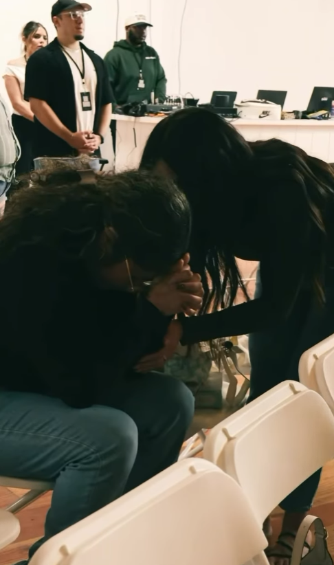 A woman is sitting with her head bowed and hands covering her face, appearing emotional or overwhelmed. Another woman is standing beside her, leaning in and holding her hand for support. In the background, several people are standing, including a man