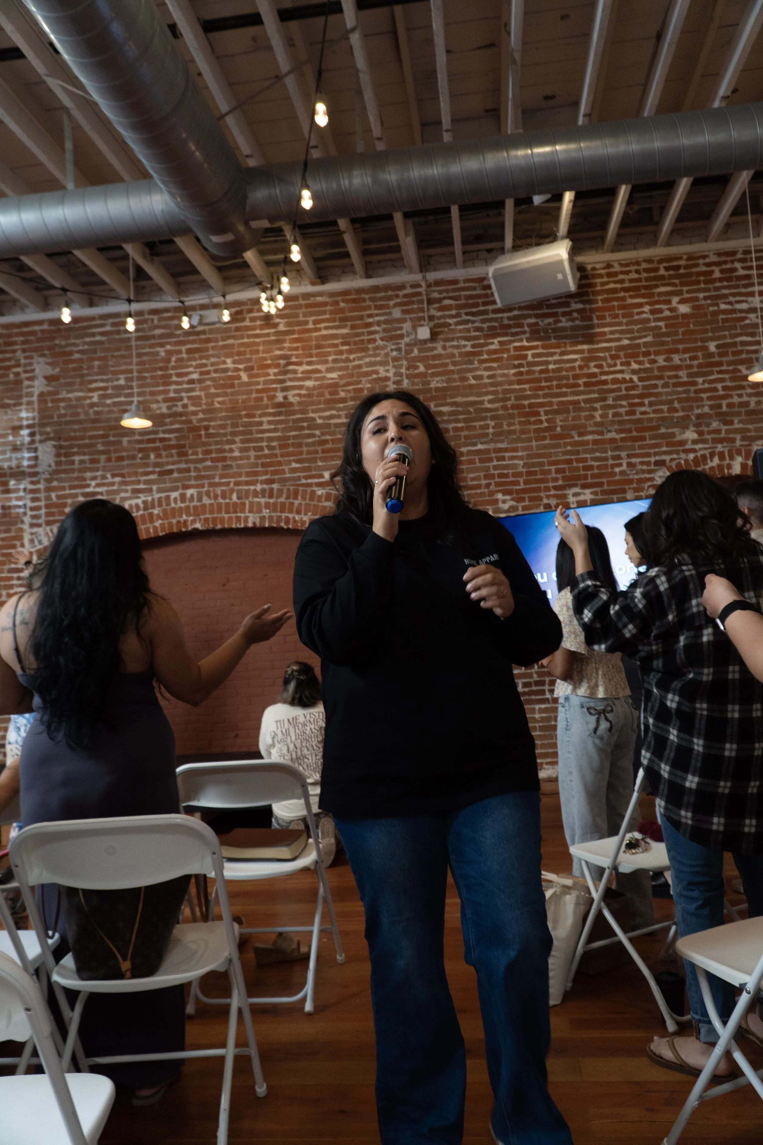 A woman singing into a microphone during a gathering in a brick-walled indoor space with string lights overhead.