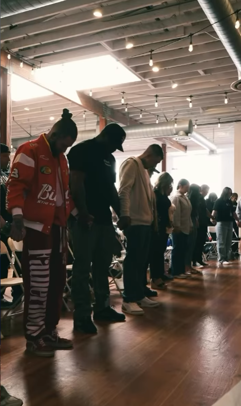 A group of people standing in a line with heads bowed in prayer inside a modern, industrial-style space with exposed ceiling beams and ductwork.