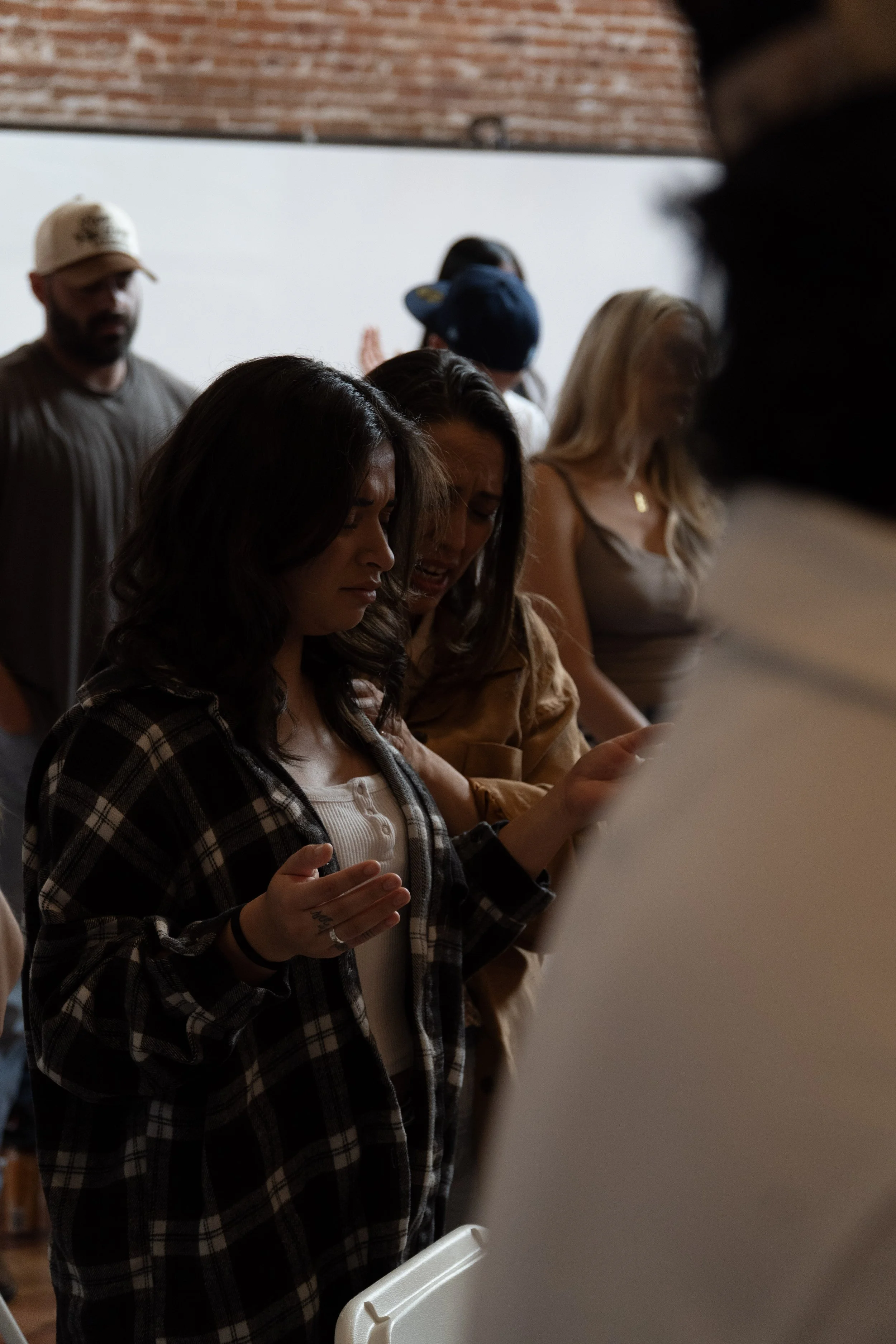 A group of diverse people in a room with brick and white walls, standing with heads bowed and eyes closed, engaged in prayer or reflection.