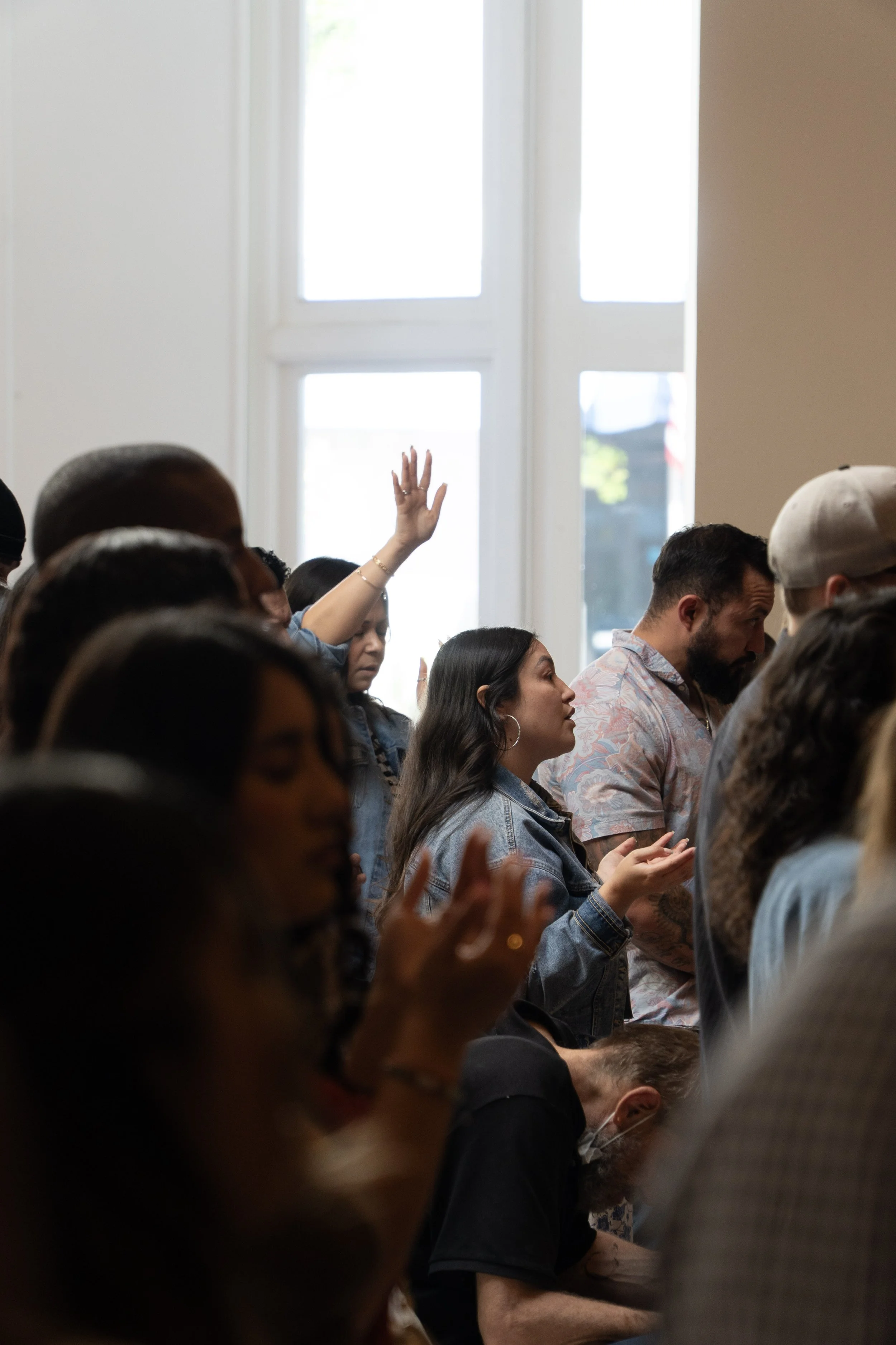 People attending a religious or community gathering in a room with large windows, one woman raising her hand.