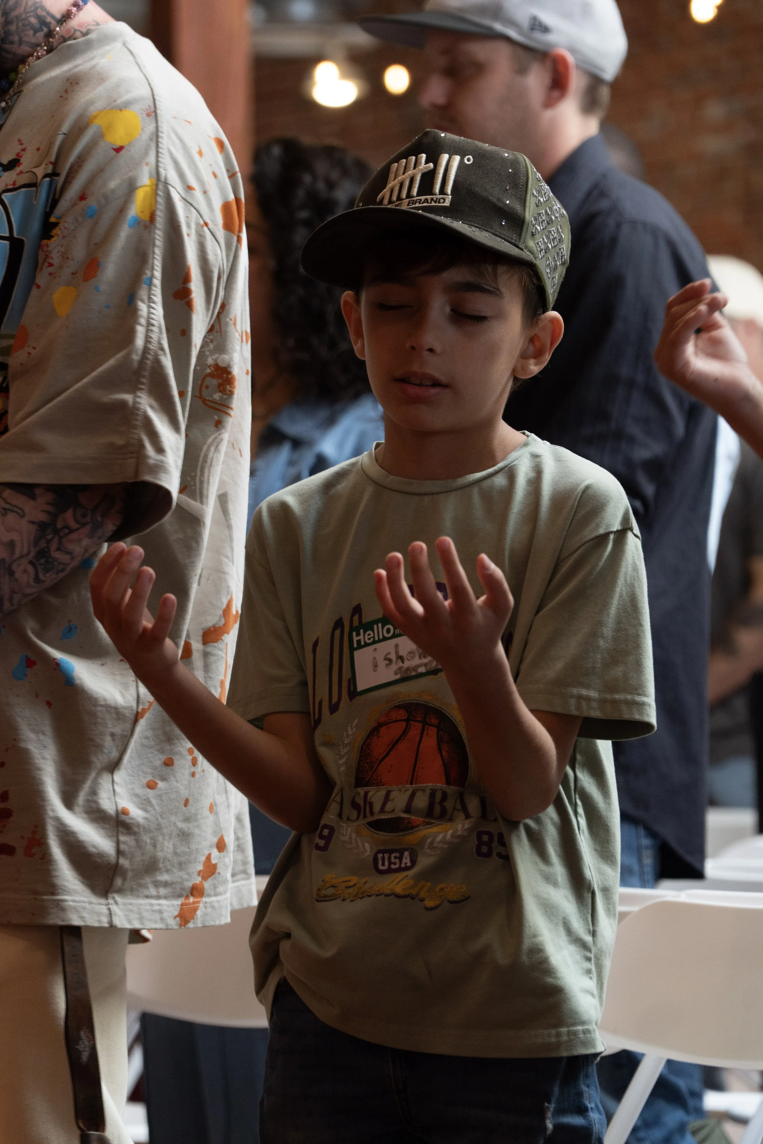 A boy in a beige T-shirt and a green baseball cap with tally marks, standing with his eyes closed and hands raised, in an indoor setting with other people in the background.