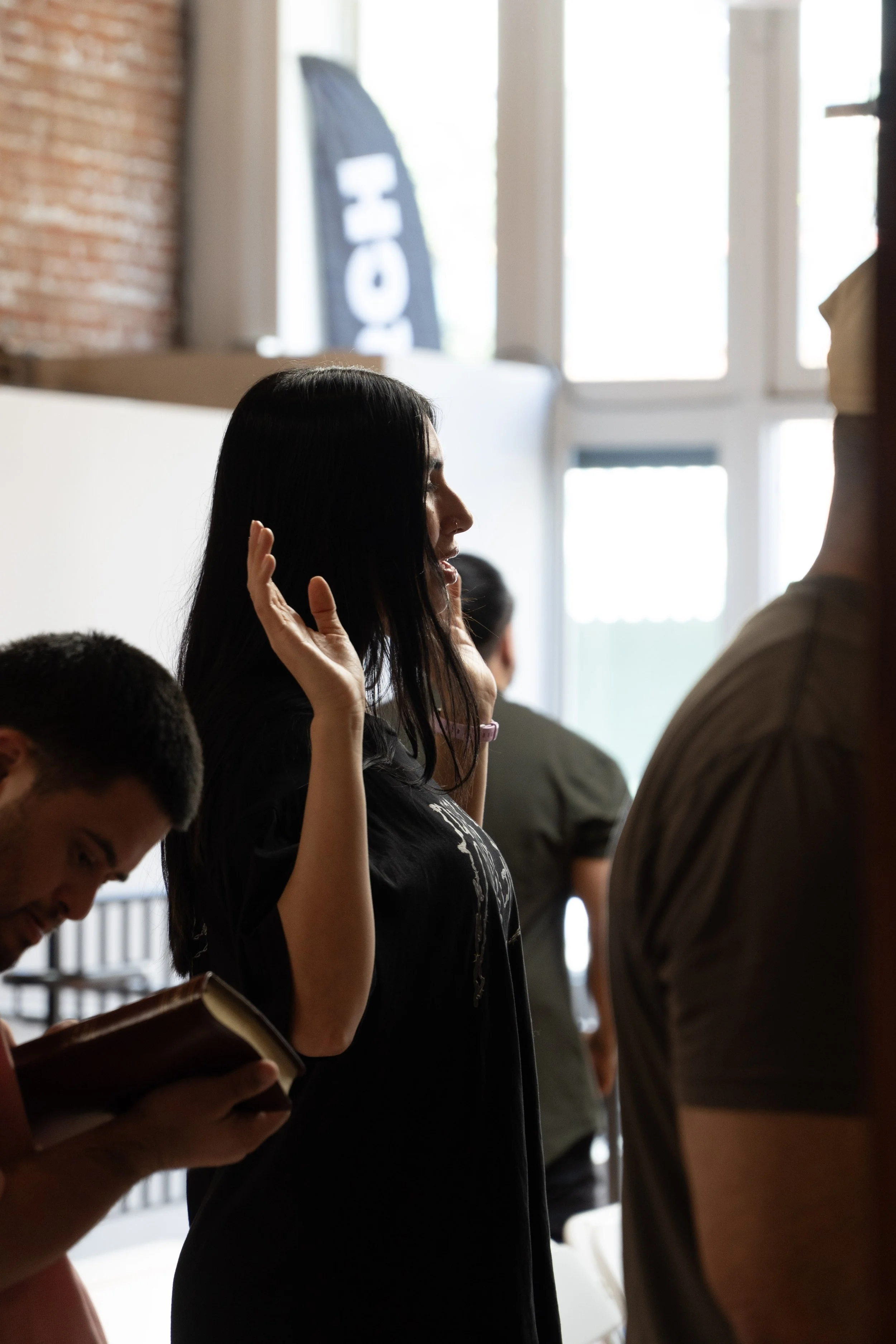 A woman with long black hair raising her hands near her face in a room with large windows and brick walls, with several other people around her, one reading a book.
