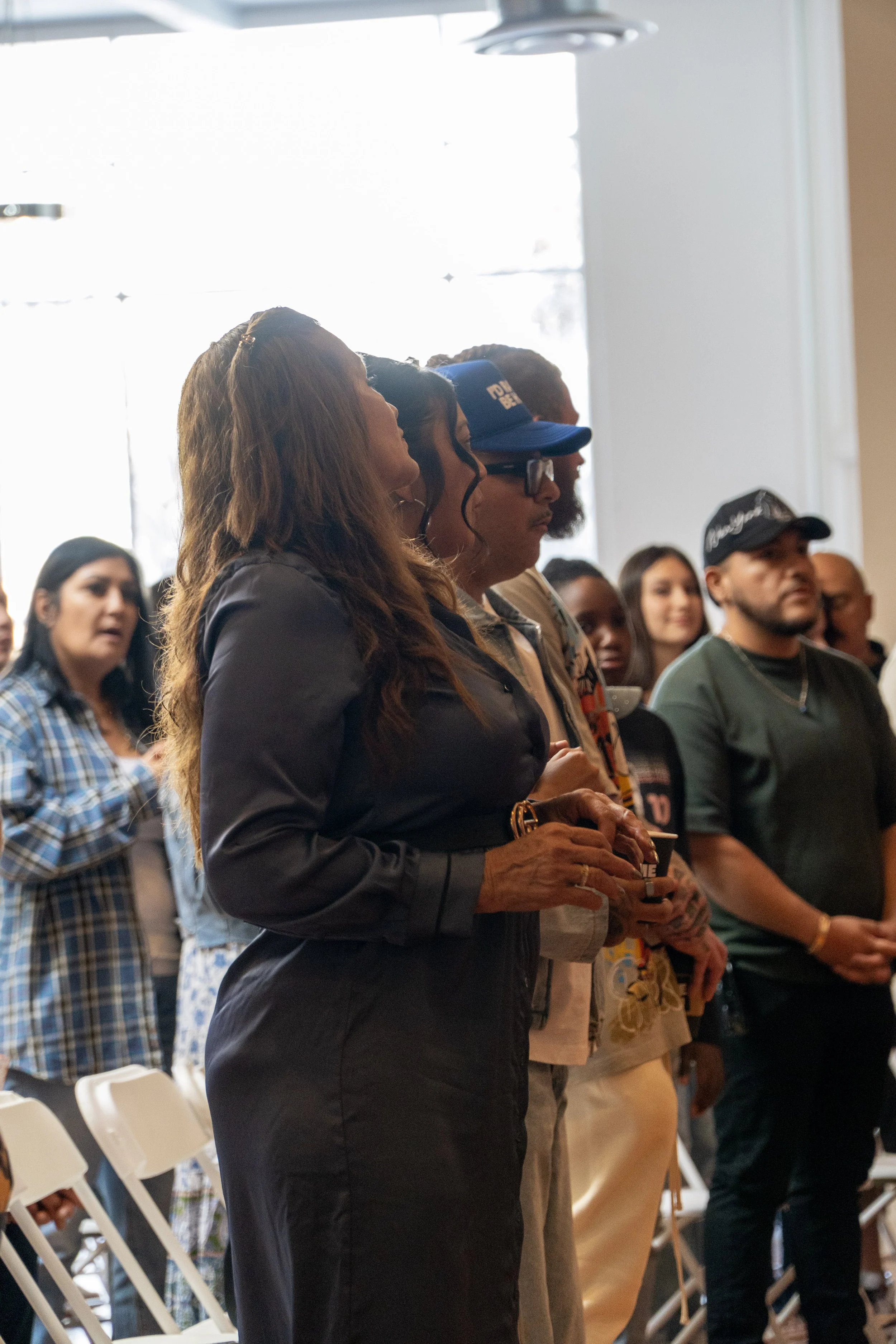 A group of diverse people standing indoors, listening attentively during an event.