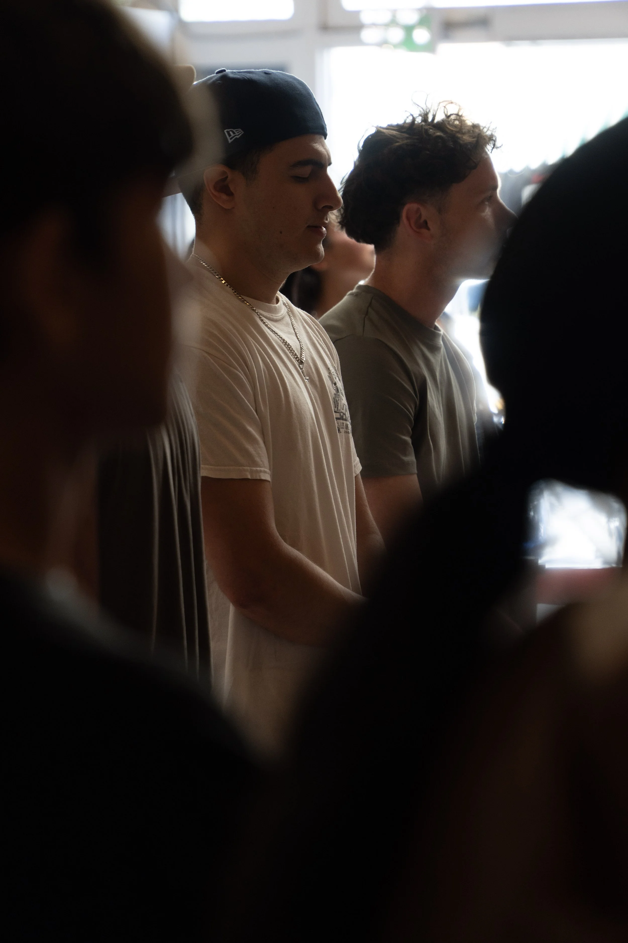 Two young men standing with eyes closed, appear to be praying or meditating, in a room with other people, natural light coming from the windows.