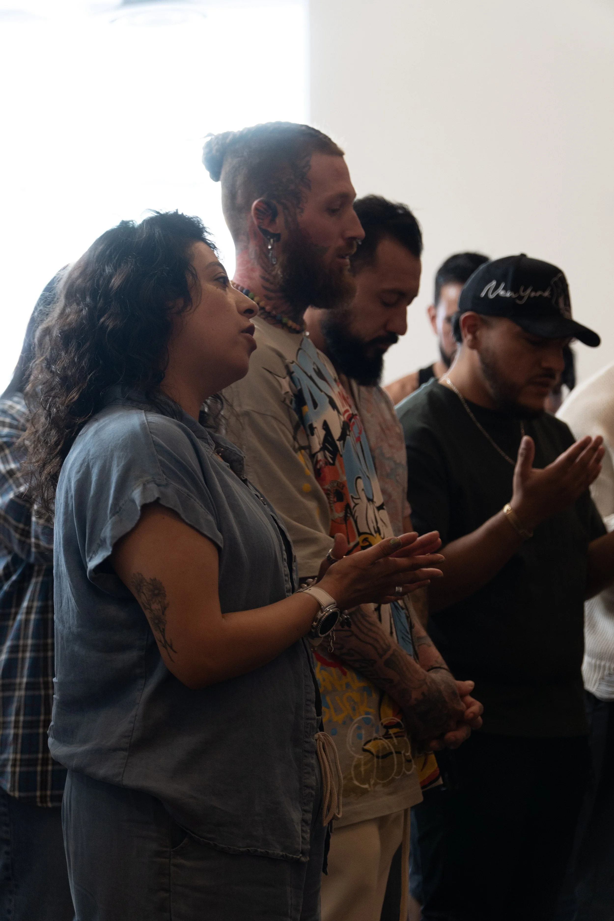 Group of people standing with eyes closed, possibly praying or meditating, in a well-lit room.