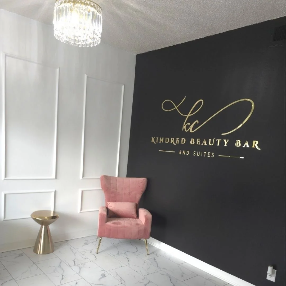Interior of a beauty salon with a black wall featuring gold lettering that reads 'KC Kindred Beauty Bar and Suites.' There is a pink velvet armchair, a small gold side table, and a chandelier hanging from the ceiling. The floor is white marble.