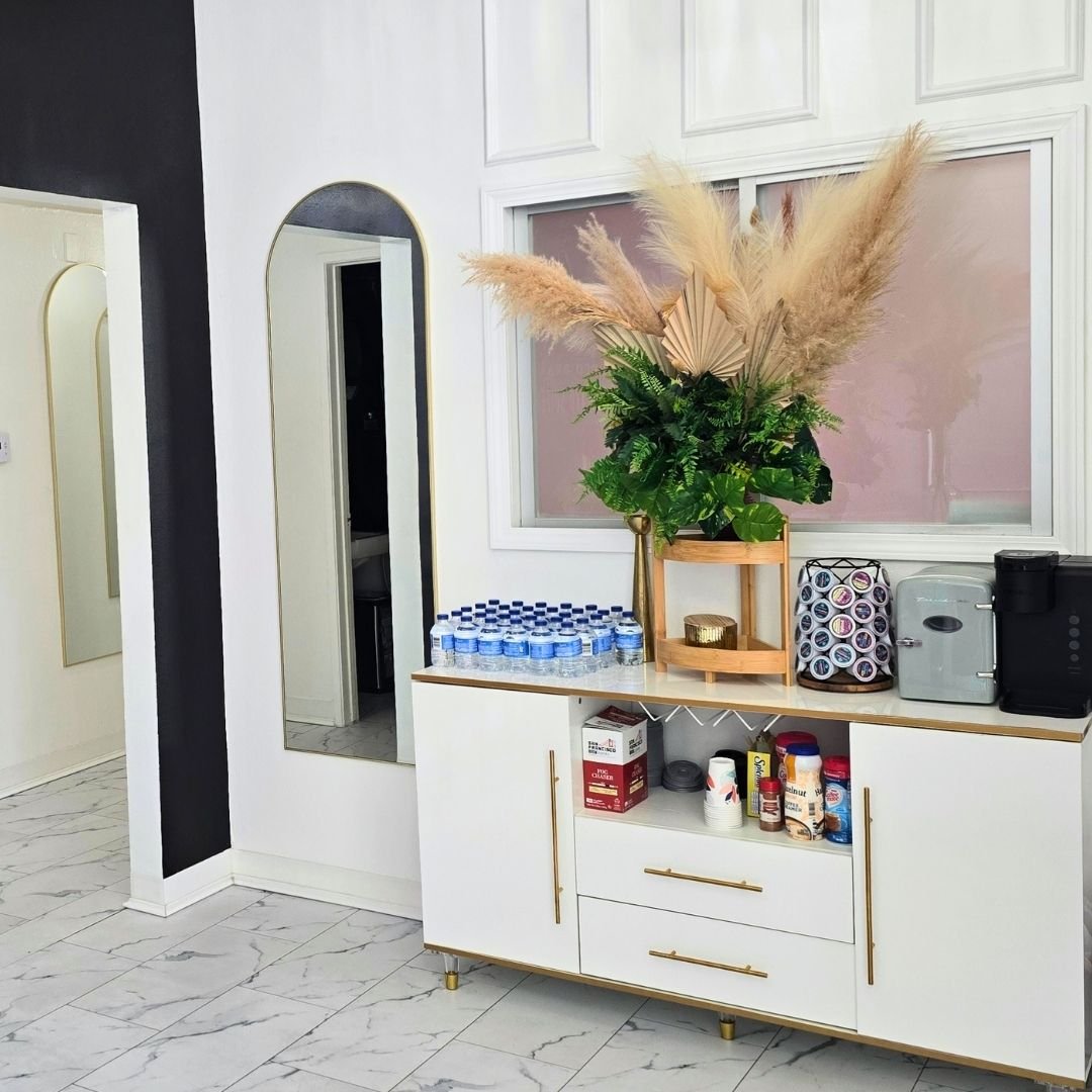 A modern interior space with a white sideboard holding bottled water, a large botanical arrangement with pampas grass, and small appliances. A mirror and a window are visible on the wall.