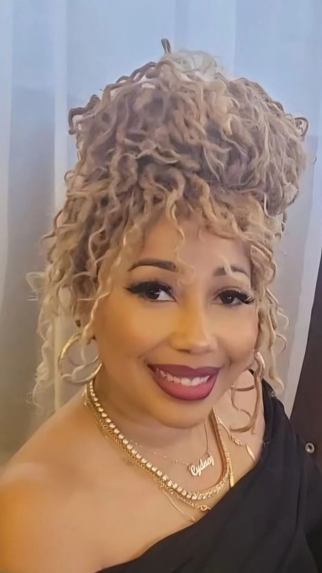 A woman with curly blonde hair styled in a large updo, smiling, wearing gold jewelry including hoop earrings and layered necklaces, wrapped in a black top or dress.