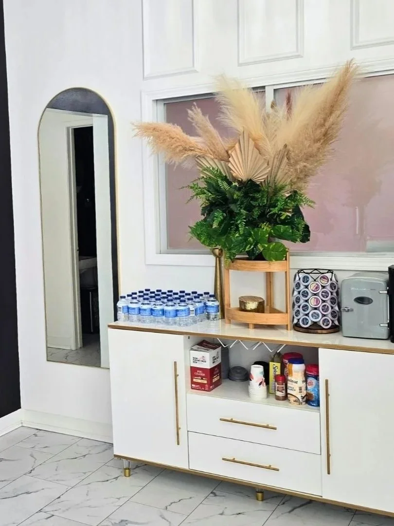 A sideboard with bottled water, a green plant with pampas grass in a wooden container, and small kitchen appliances, in a modern interior with marble floor and white panel walls.