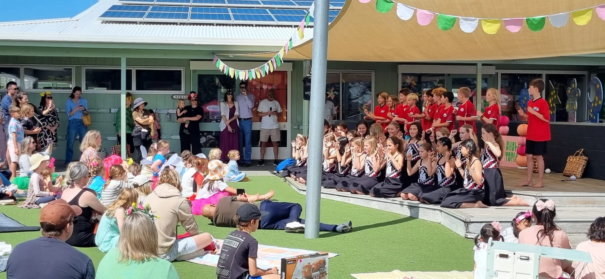 Children and adults attending an outdoor event, with children performing on a stage dressed in black and red costumes, some in red shirts and others in black and white patterned outfits. The audience, including some wearing hats and costumes, is seat