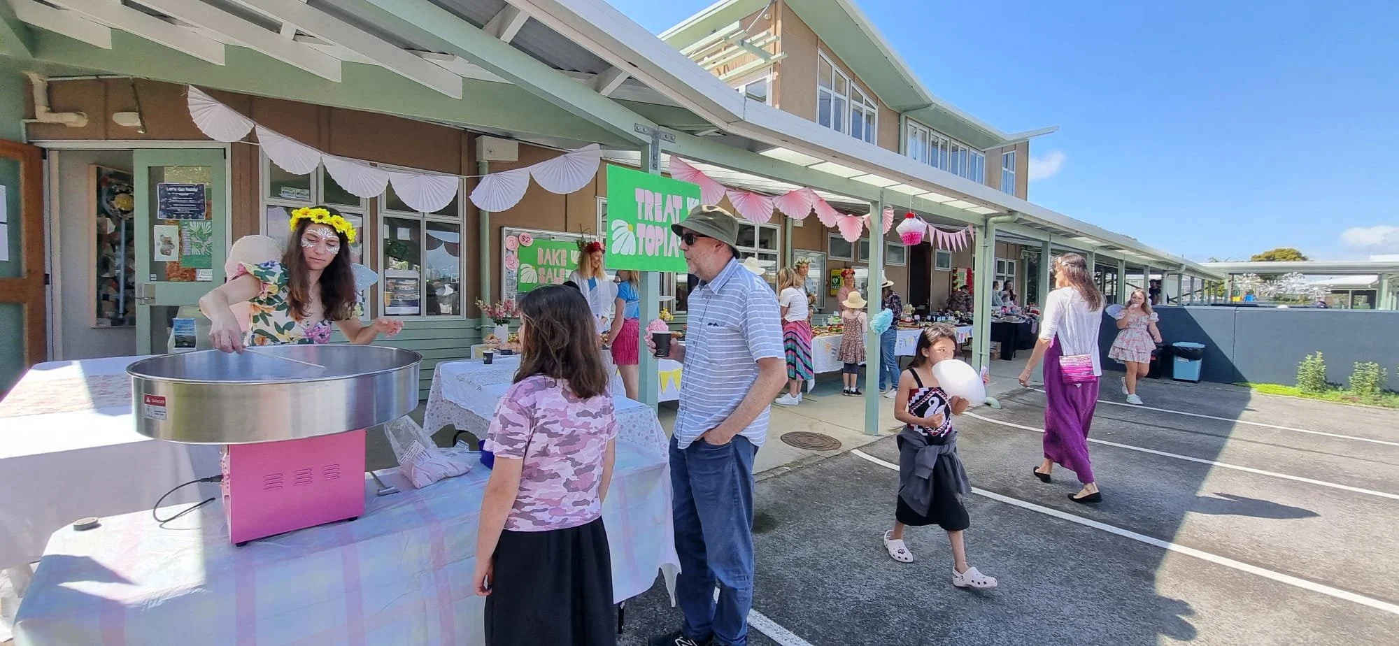 People at an outdoor market stand decorated with pink and blue paper banners, selling baked goods and crafts on a sunny day.