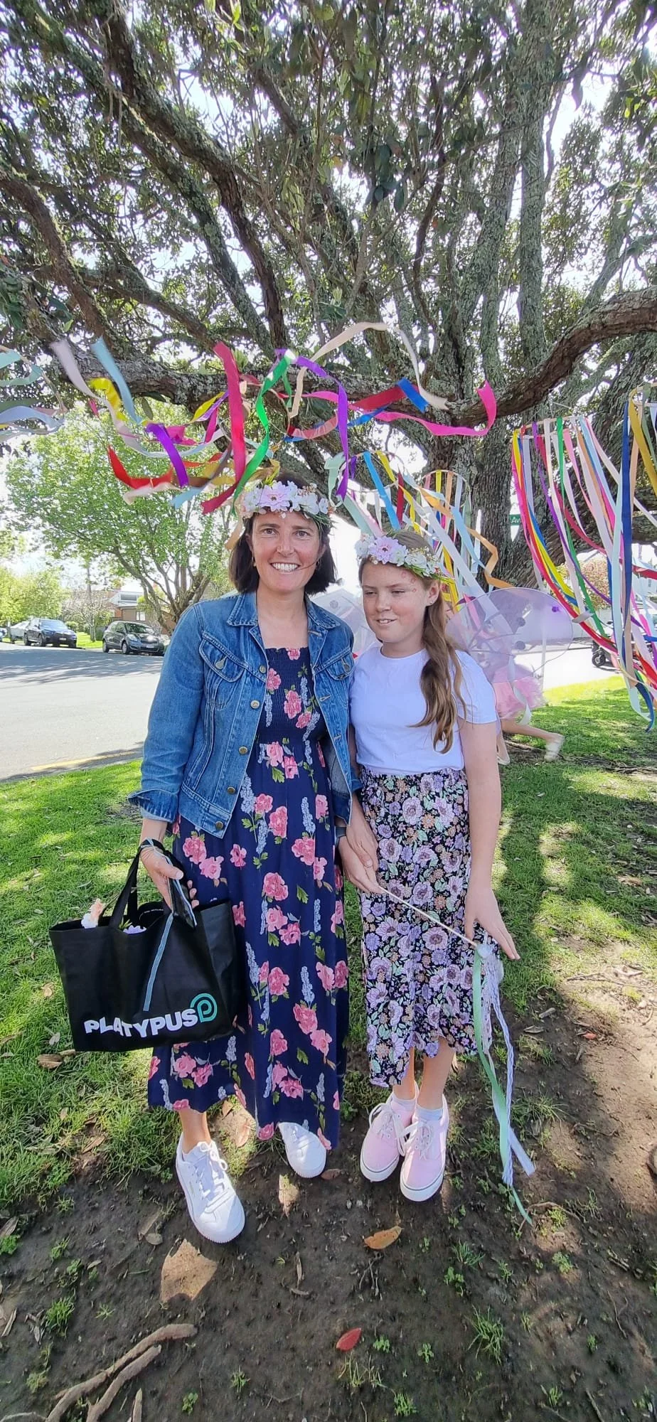 A woman and a girl wearing flower crown headbands celebrate under a tree decorated with colorful streamers. The woman holds a black tote bag with "PLATYPUS" written on it, and the girl carries a wand. They are smiling on a sunny day in a park.