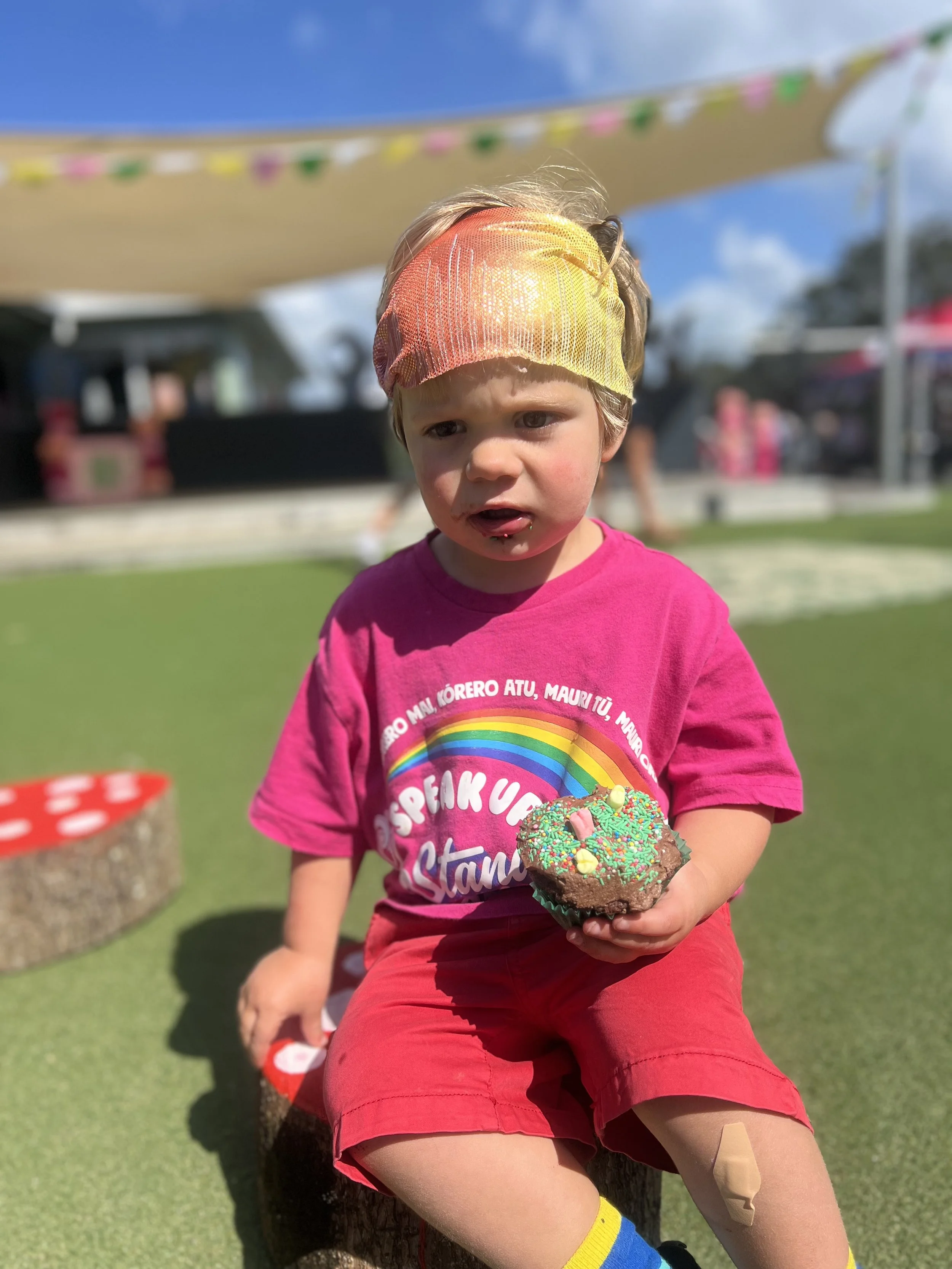 A young boy with a colorful headband and colorful T-shirt sitting on a tree stump outdoors, holding a decorated chocolate cupcake with green sprinkles and small candy toppings, with a casual and festive outdoor setting in the background.