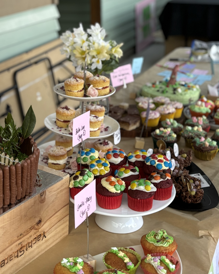 Display of decorated cupcakes and mini cakes with flowers on a table, with handwritten notes and pink sticky notes.
