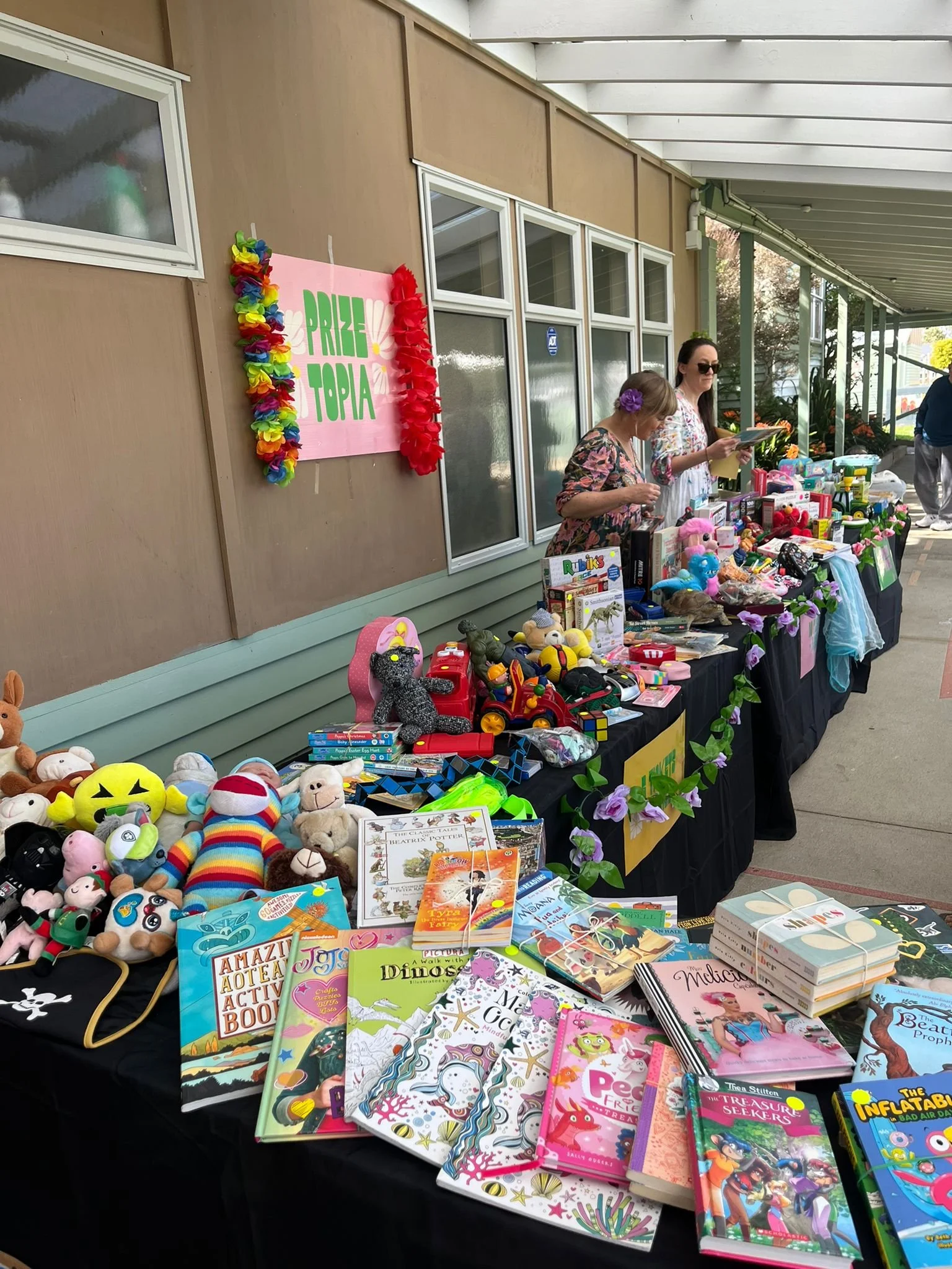 A table at a yard sale or fair with toys, books, and games, under a canopy outside a building, with two women behind it, and a pink sign that reads 'Prize Topia' decorated with colorful leis.