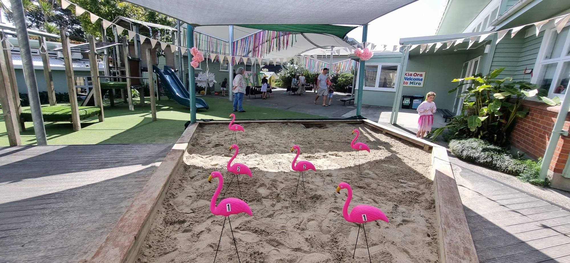 Sandpit with pink flamingo-shaped yard decorations numbered 1 to 5, children playing and people gathering, decorated for a celebration at a playground with a slide, shaded area, and building with a sign that reads 'Kia Ora Welcome to Miro'.