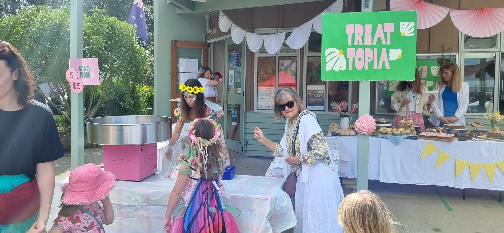 People gathered at an outdoor event with a table of sweets and a green sign that says "Treat Topia." There are children and adults, some wearing flower crowns or butterfly wings, and a stall in the background with pink and yellow decorations.