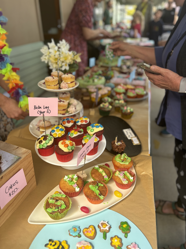 Display of decorated cupcakes at a bakery or market stall, with colorful icing and sprinkles, and small signs labeling them.