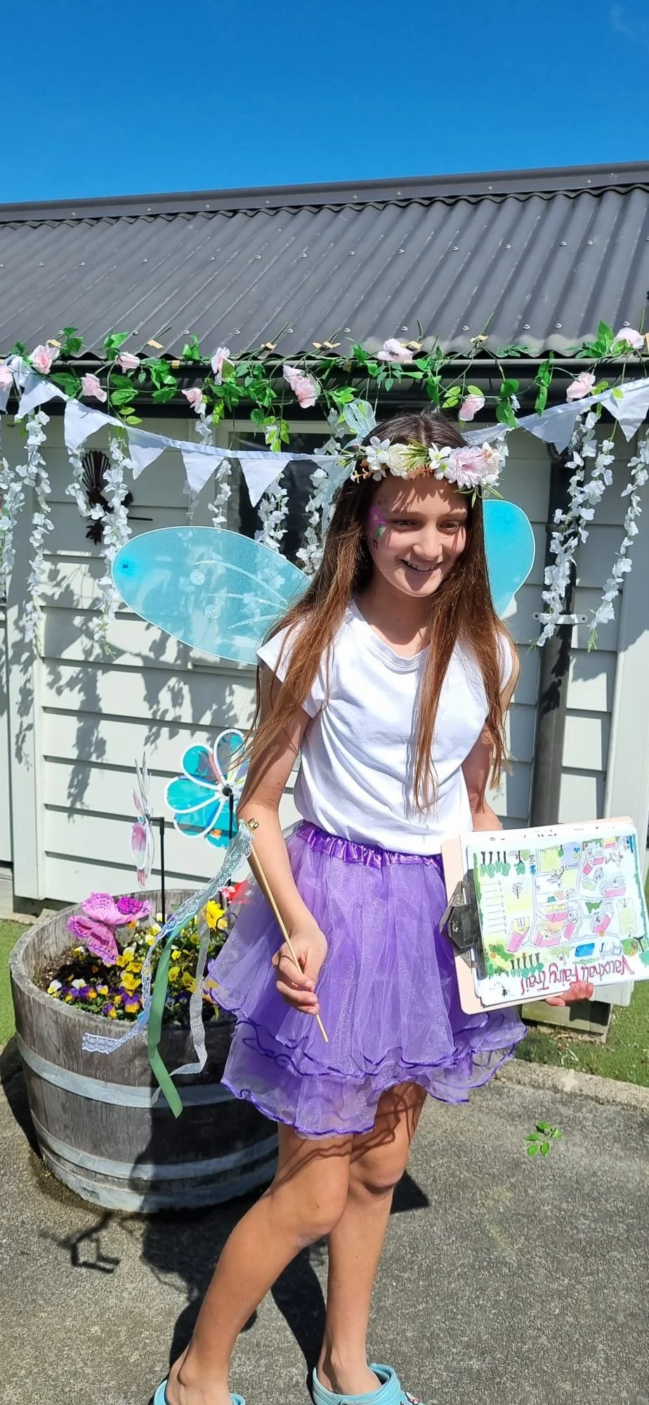 A young girl dressed as a fairy with blue wings, a purple tutu, and a flower crown, standing outdoors in front of a decorated backdrop with flowers and bunting, holding a wand and a drawing tablet, smiling.