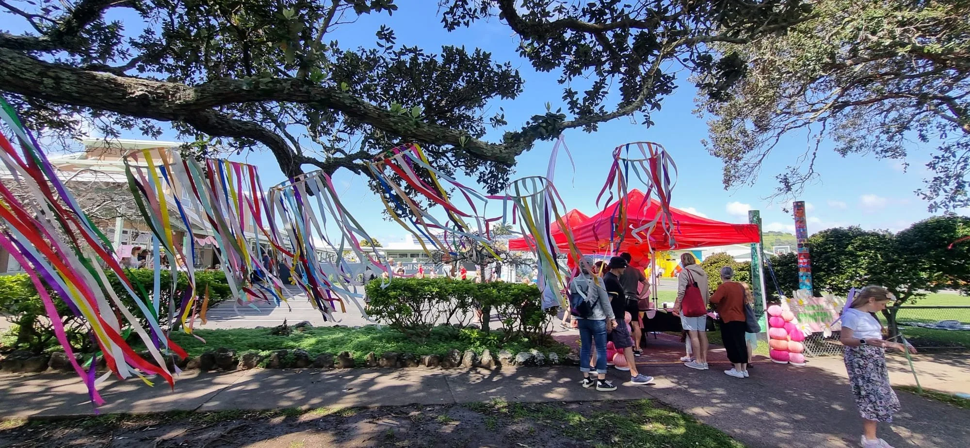Outdoor community event with colorful streamers hanging from a tree, a red tent, and people gathered around tables on a sunny day.