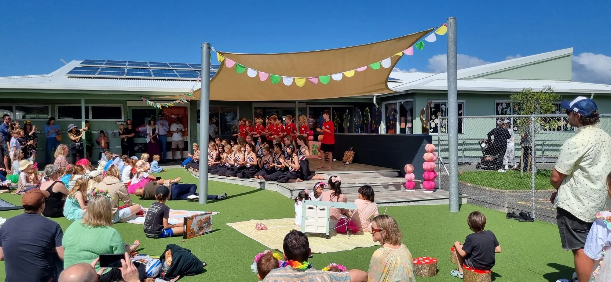 Children and adults attending an outdoor performance at a school or community event, with a stage decorated with colorful bunting and pink paper lanterns, on a sunny day.