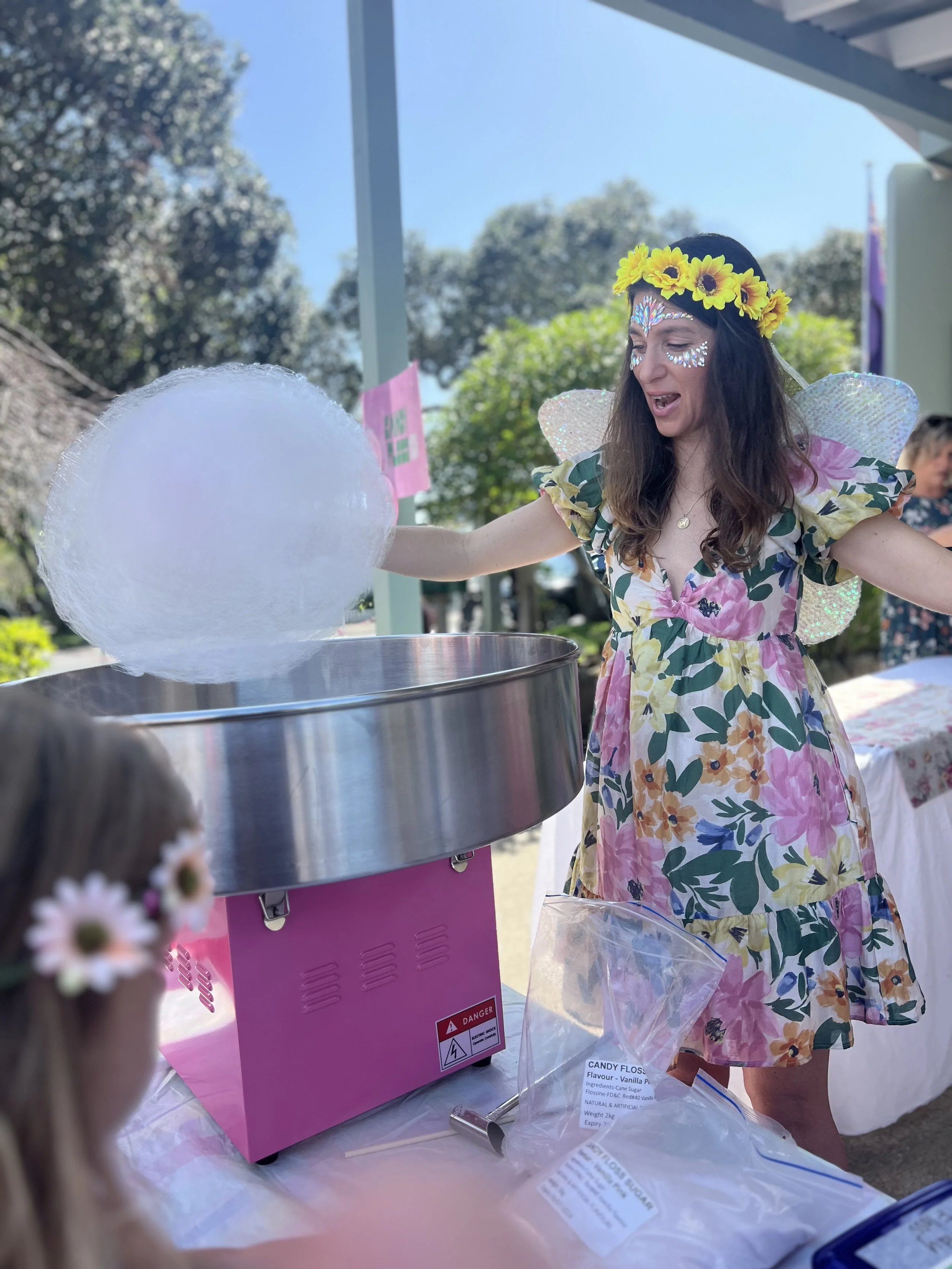 A woman dressed as a fairy with a yellow flower crown and sparkly face decorations makes cotton candy at an outdoor event. She is wearing a floral dress with puffy sleeves and has fairy wings on her back. A young girl with a floral headband watches h