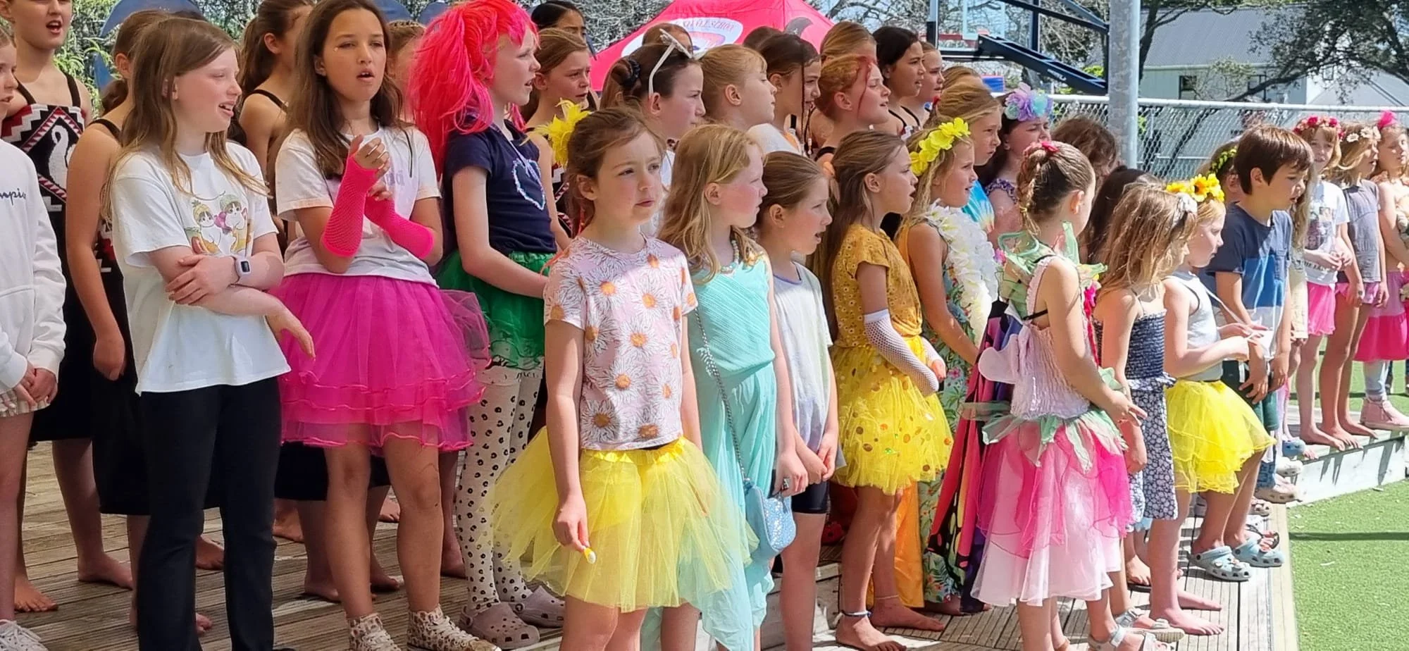 Group of young children dressed in colorful costumes, standing outdoors during daytime, participating in a school performance or event.
