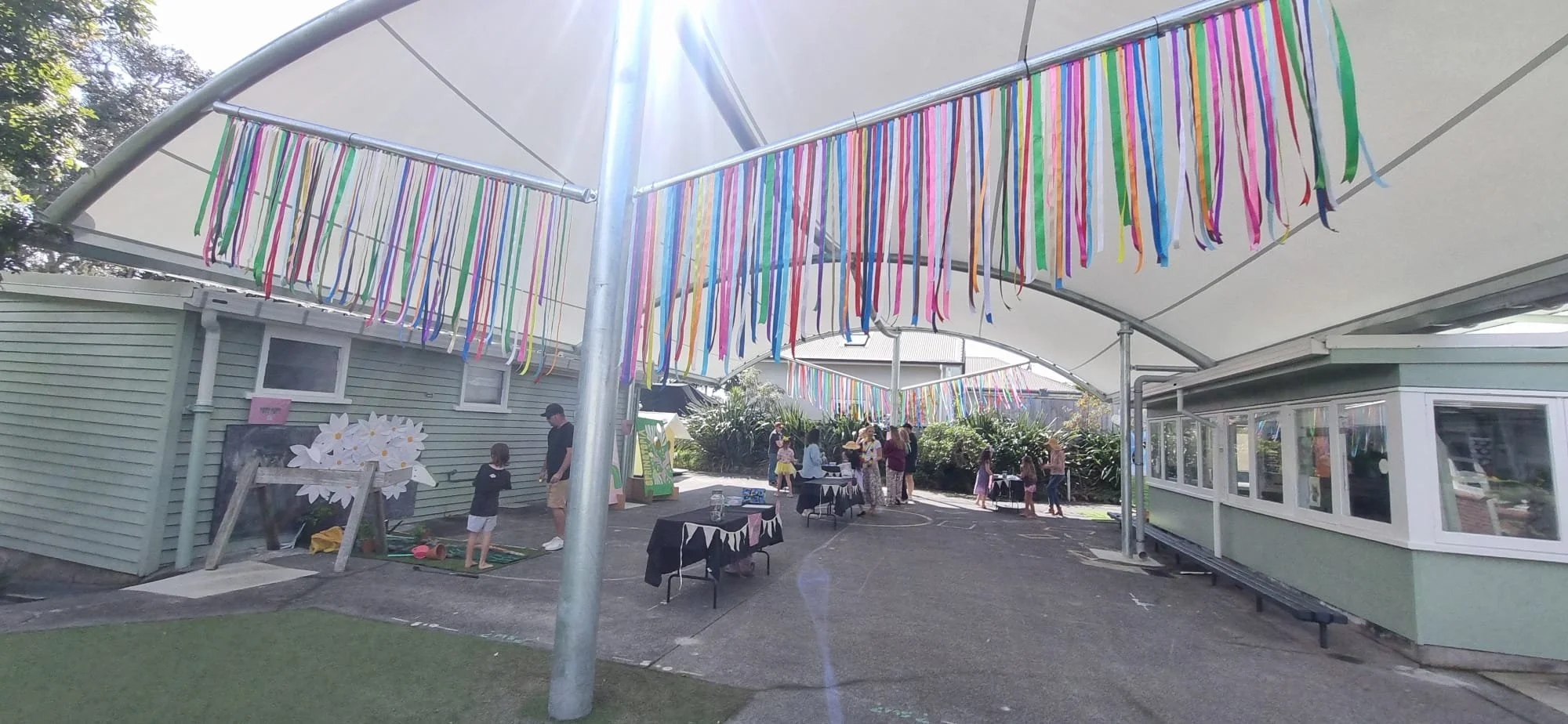 Outdoor community event with colorful streamers hanging overhead, tables with black tablecloths, and people including children and adults gathering in a garden area.