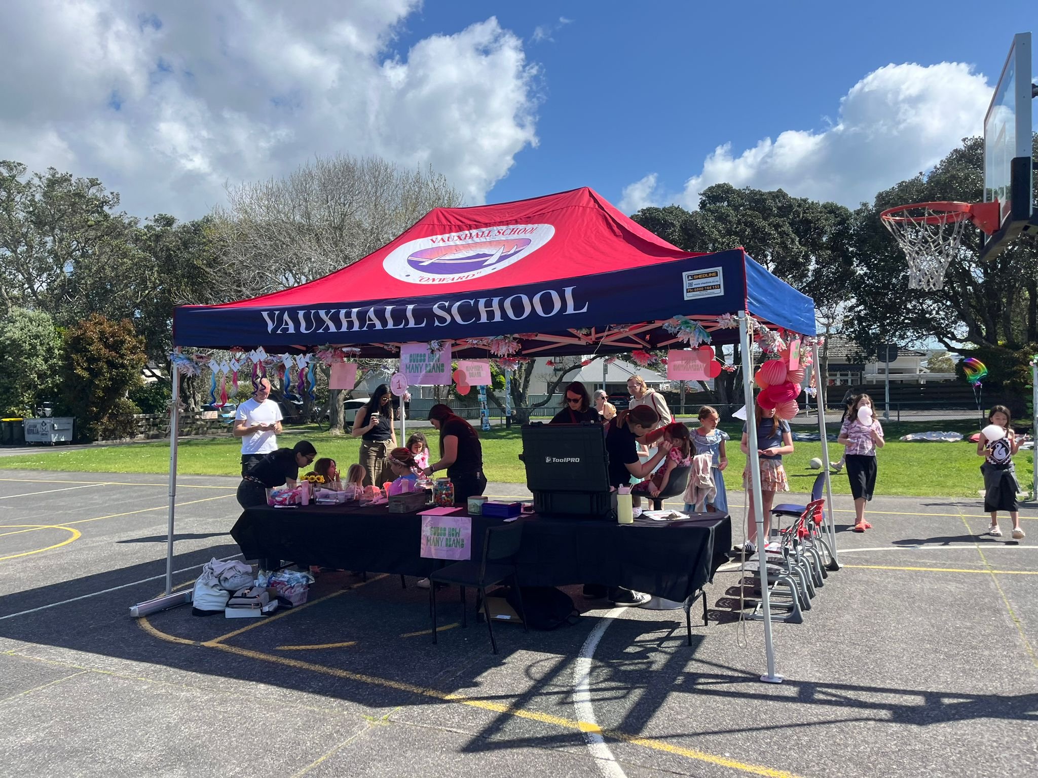 A school event takes place outdoors on a basketball court, with a red and blue tent labeled 'Vauxhall School.' Several children and adults are gathered around tables decorated with pink and purple balloons and banners, participating in activities. Th