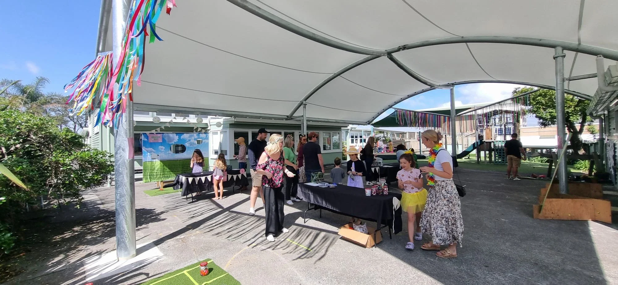 Children and adults at an outdoor celebration under a large canopy, with tables displaying items, colorful banners, and a playground in the background.