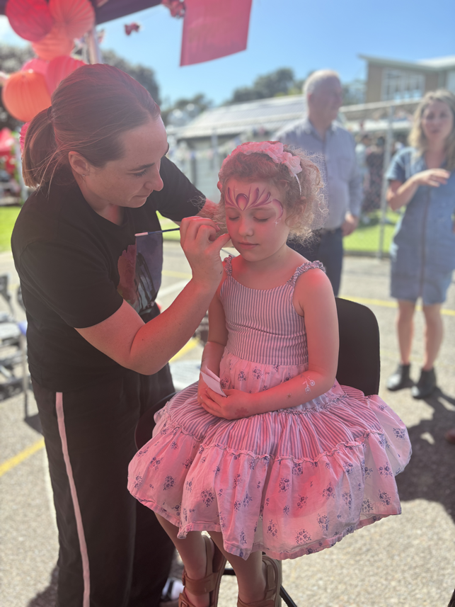 A young girl sitting on a chair getting her face painted with a pink, purple, and white design by a woman at an outdoor event. The girl is wearing a pink layered skirt with floral patterns and holding a white paper. In the background, there are a few