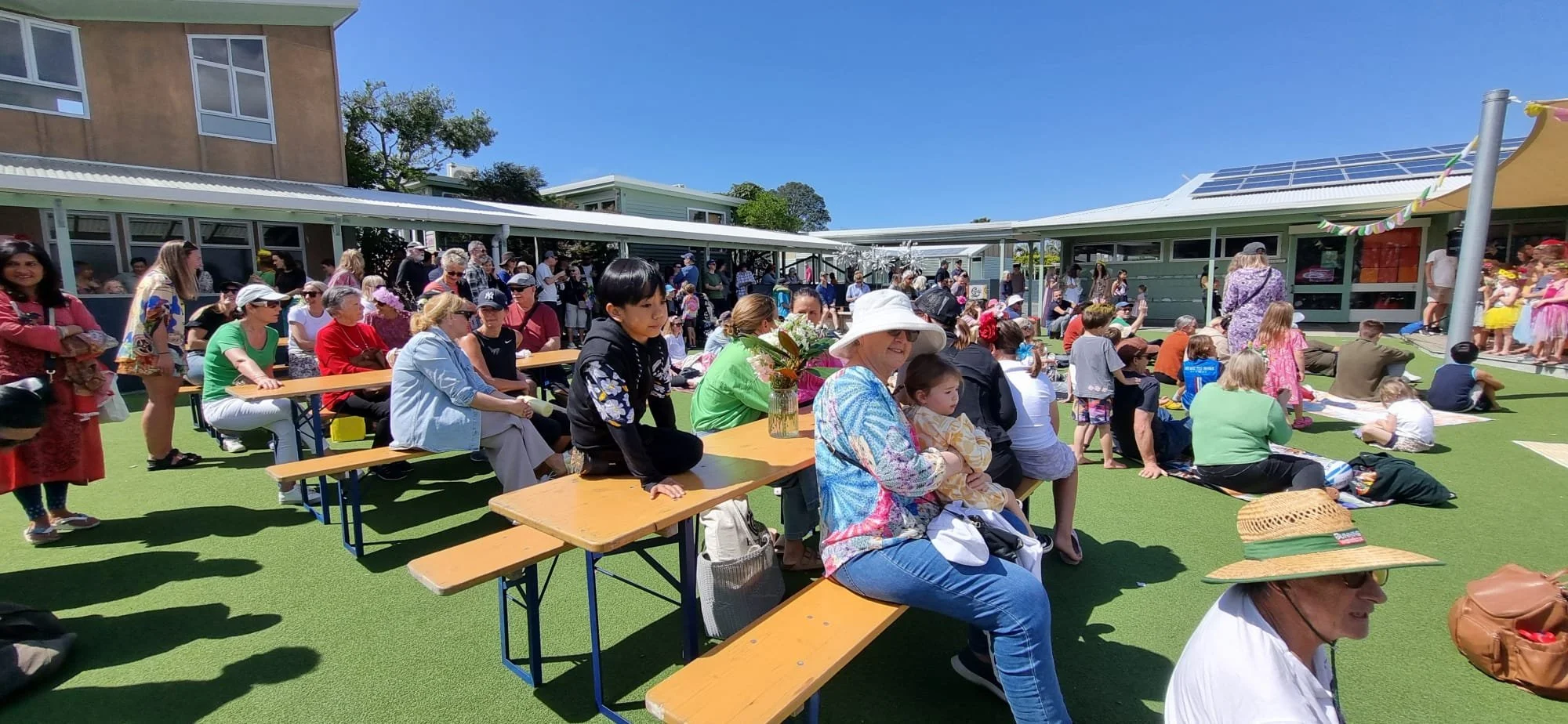 A diverse group of people gathered outdoors at a daytime event, sitting and standing on artificial green turf, with children on a small stage and adults sitting at picnic tables. The scene is sunny with a blue sky, and there are buildings with solar 