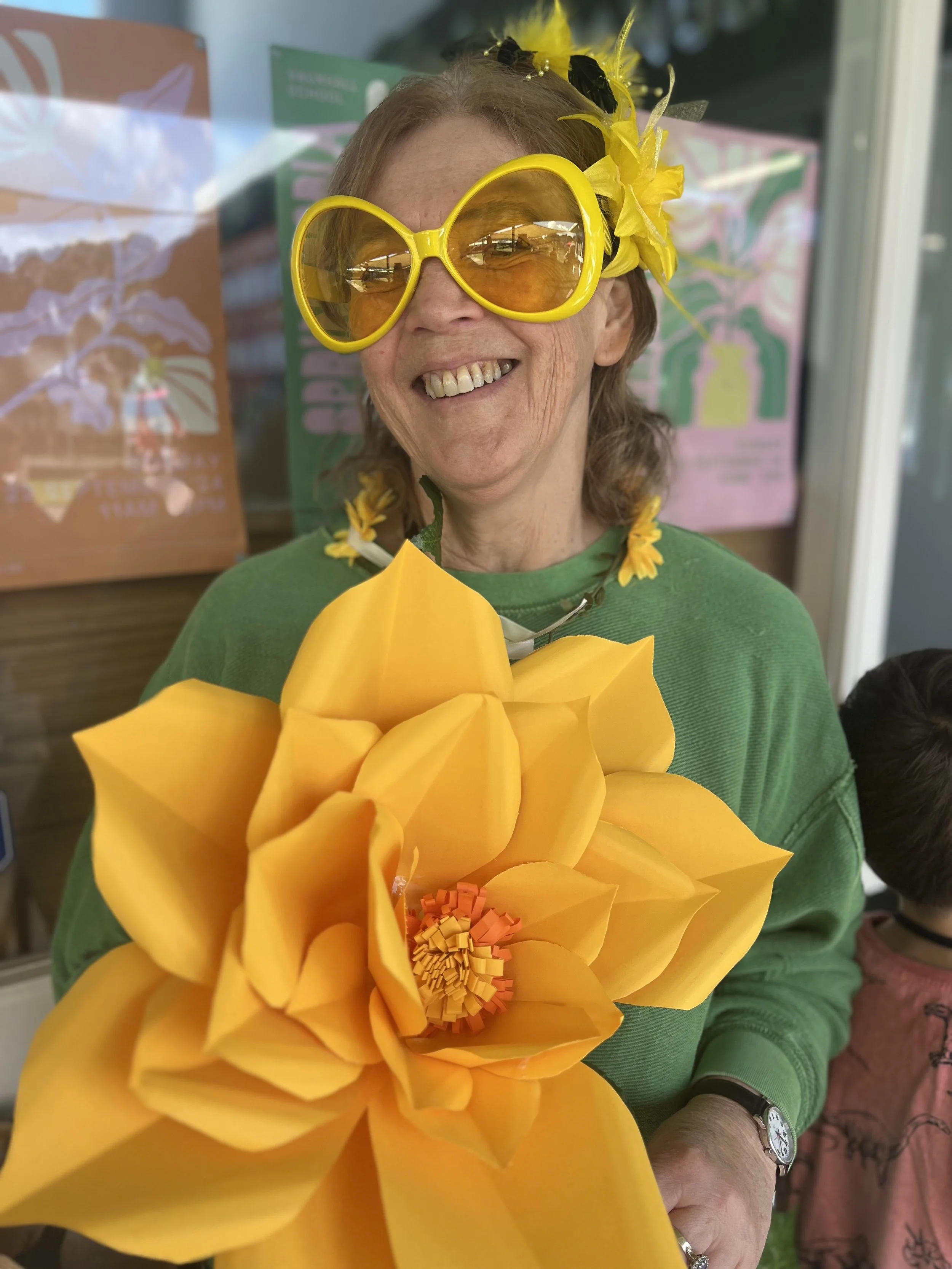 A woman wearing large yellow sunglasses, a yellow flower headband, and a green shirt, smiling and holding a large paper flower. The background features colorful posters and a window.