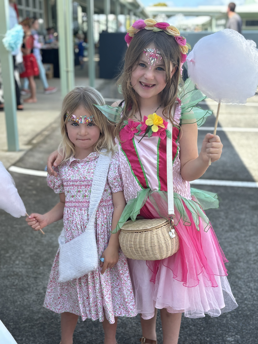 Two young girls dressed in fairy costumes at an outdoor event, with face paint and face gems, one holding cotton candy.