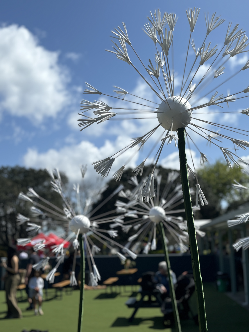 Close-up of decorative dandelion-shaped flowers with white petals on green stems against a blue sky with clouds, at an outdoor gathering.