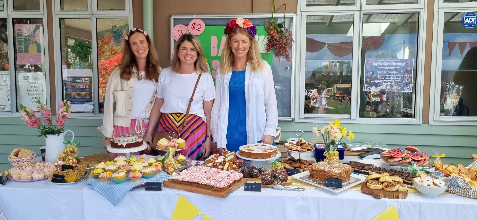 Three women standing behind a table filled with assorted baked goods at an outdoor event, with a green sign and windows in the background.
