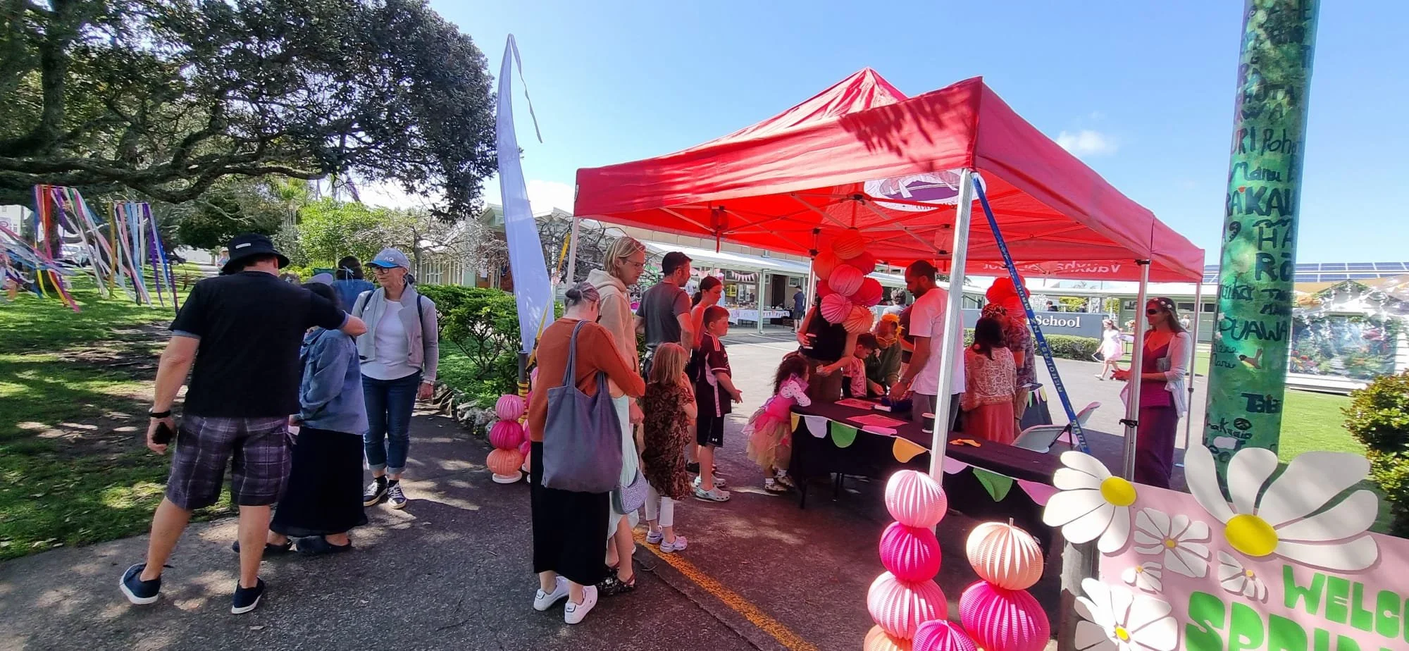 People gathered at a booth decorated with pink paper lanterns and flowers at an outdoor event on a sunny day.
