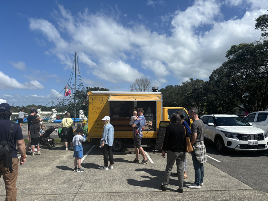 People gather around a yellow food truck in a parking lot near a playground with a climbing structure, with cars parked nearby and trees in the background under a partly cloudy sky.