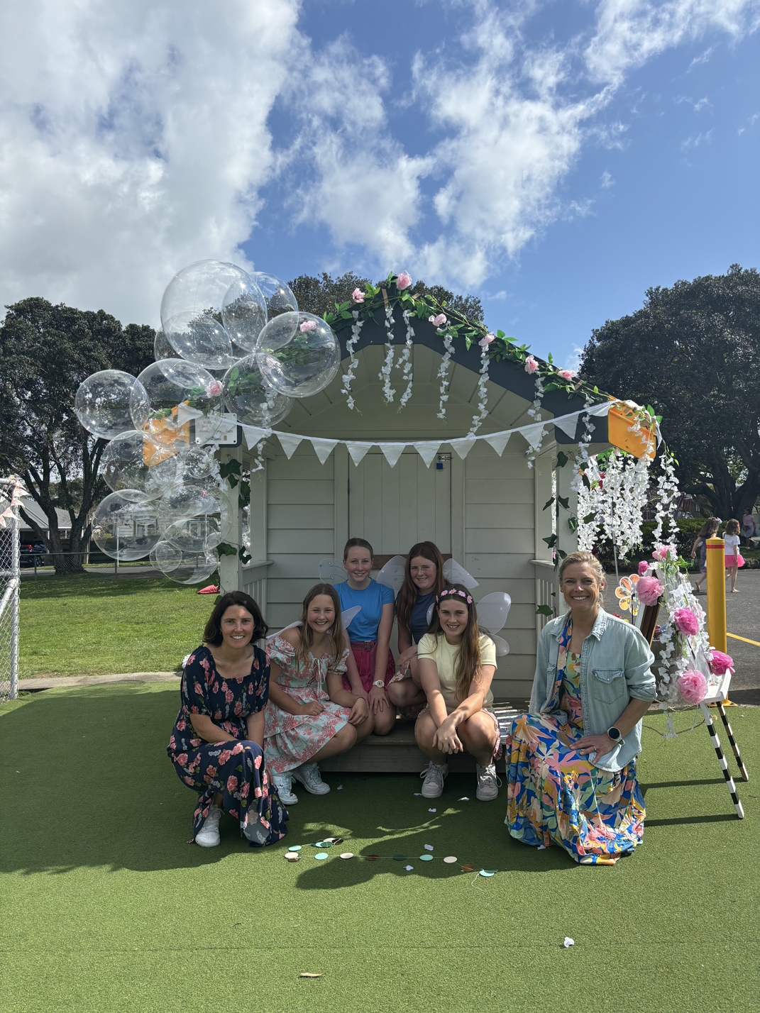 Group of six women and girls dressed as fairies posing in front of a decorated fairy garden shed at an outdoor event on a sunny day.