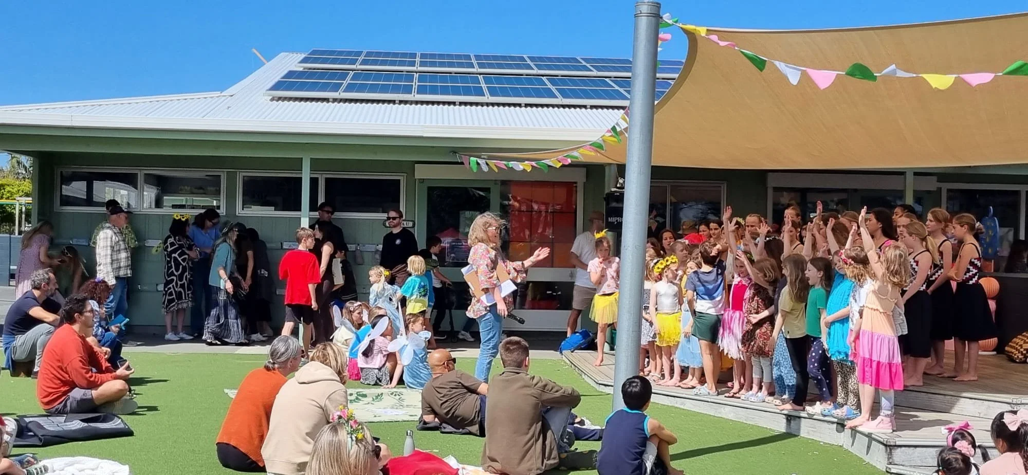 Children and adults gathered outside for a group activity, with children dressed in colorful costumes and some wearing fairy wings, while others are standing on a stage and a woman appears to be leading or conducting the activity.