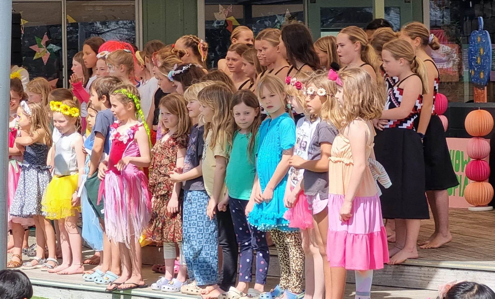 Group of children standing on outdoor stage in colorful casual and festive clothing, preparing for a performance.