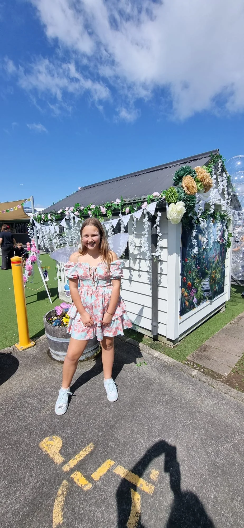A young girl dressed as a fairy with white fairy wings, a floral dress, standing on a parking lot, smiling next to a decorated small white building with flowers and fairy lights.