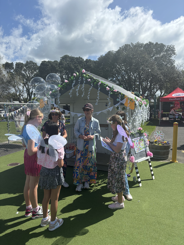 Children and adults gathered around a decorated booth with balloons and flowers at an outdoor event on a sunny day.