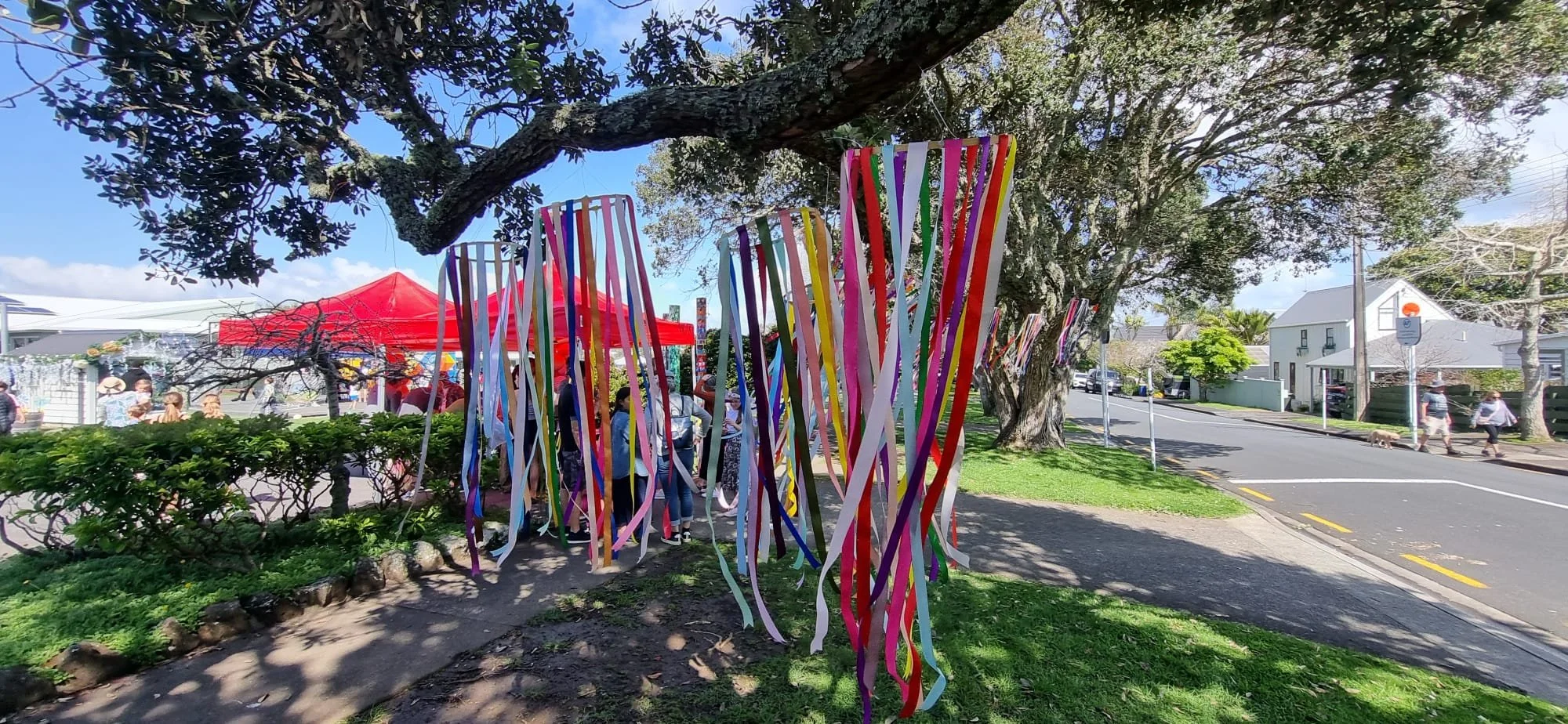 Colorful streamers hanging from a tree at an outdoor event with tents and people gathered underneath. Residential street nearby with pedestrians and parked cars.