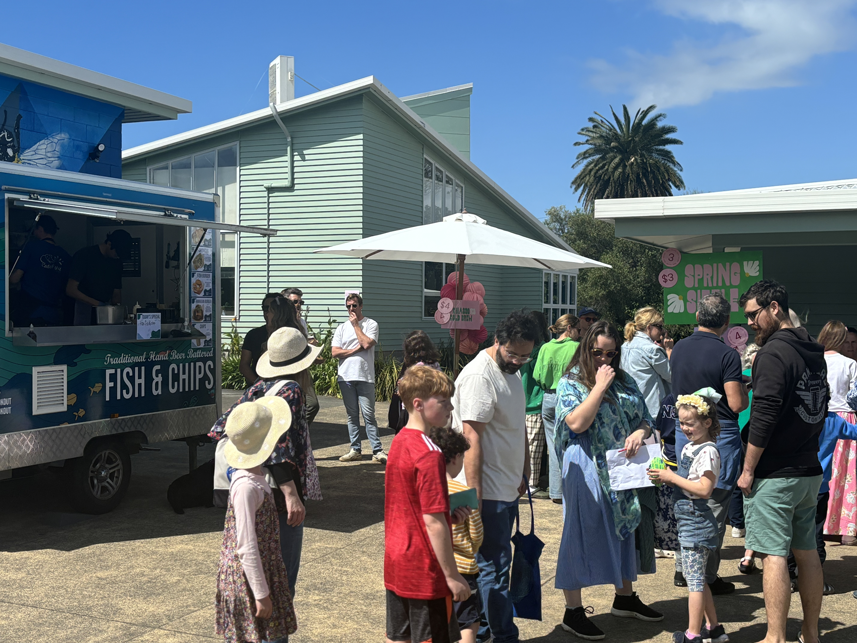 People standing in line at a food truck selling fish and chips during daytime at an outdoor event with a pastel green building and a palm tree in the background.