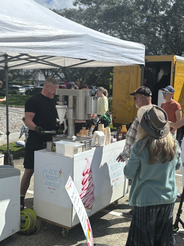 A man preparing and serving ice cream at a mobile ice cream stand under a white canopy. Customers are in line, with a woman wearing a hat and glasses in the foreground. The stand has a sign that reads 