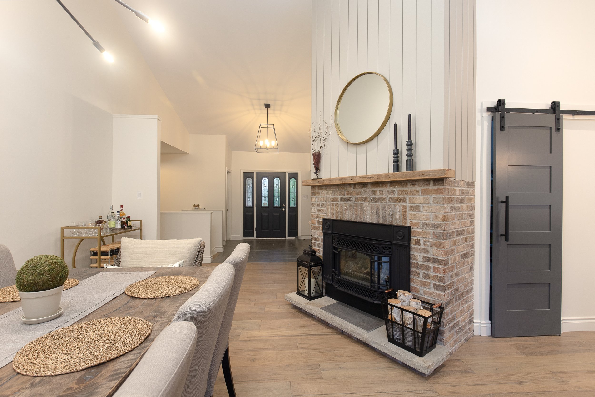 Living room with a brick fireplace, wooden mantel, circular mirror, black sliding barn door, dining table with placemats, and a hallway with front door in the background.